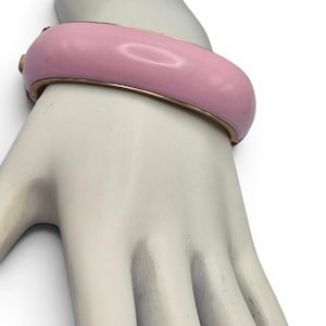 Könnte beinhalten: Ein hellrosa Armreif mit goldfarbenen Rändern. Das Armband hat eine abgerundete Form und scheint aus Emaille oder einem ähnlichen Material zu bestehen. Das Armband wird an einer Mannequin-Hand gezeigt.