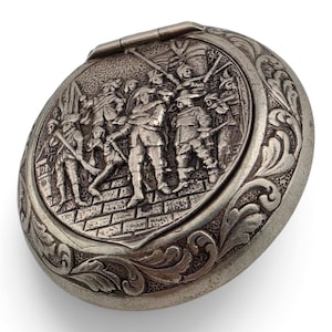 Puede incluir: Una caja redonda de metal plateado con tapa abatible que presenta un relieve detallado de un grupo de figuras con vestimenta histórica sobre una superficie pavimentada. El borde exterior de la tapa está decorado con un patrón de hojas en relieve.