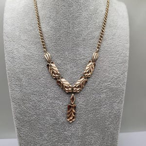 May include: A gold-toned necklace with a delicate floral design. The necklace features a chain and a pendant with intricate leaf and flower details. The pendant hangs from a series of stylized leaves. The necklace is displayed on a gray mannequin.