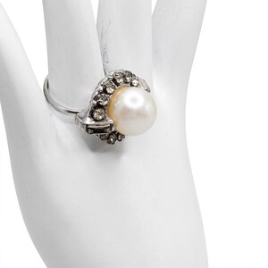 May include: A silver-toned ring featuring a central round white pearl surrounded by a cluster of small clear faceted stones. The setting includes a mix of round and baguette-shaped stones arranged in a decorative pattern around the pearl.