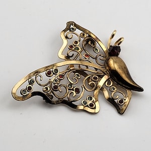 May include: A gold-toned butterfly brooch with intricate filigree wings. The wings are adorned with red, green, and clear gemstones. The butterfly's body is a darker gold color, and the antennae are visible.