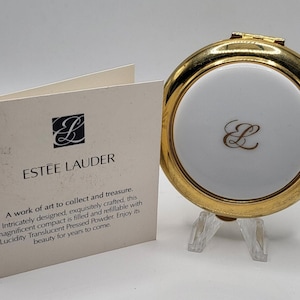 May include: A gold-toned compact with a white center and a gold 'L' emblem, next to a card with the Estée Lauder logo and text. The compact is displayed on a clear stand, showcasing its design.