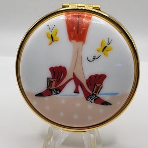 May include: A round, gold-toned compact mirror with a painted design. The design features a woman's legs and feet in red high heels, with yellow butterflies on a white background. The compact is displayed on a clear stand.