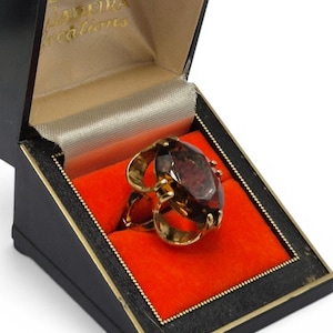 May include: A gold-toned ring with a large, oval, brown gemstone, presented in a black jewelry box with a red interior. The box is open, revealing the ring's intricate design and the brand name "LADOIRA creations".