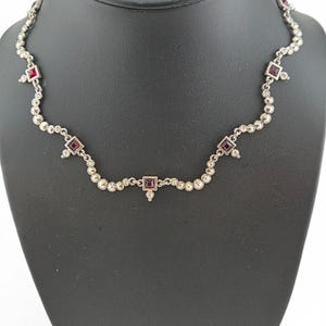 May include: A silver-toned necklace featuring a delicate chain adorned with small, round, metallic beads and four evenly spaced square-cut, deep red gemstones. Each gemstone is set in a square frame with a small dangling bead at the bottom.