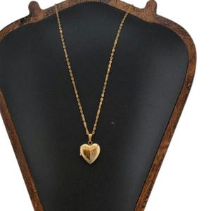 May include: A gold-toned metal necklace featuring a delicate chain and a heart-shaped locket pendant. The locket has a textured surface with a split design, showing a smooth polished section on one side and an engraved floral pattern on the other.