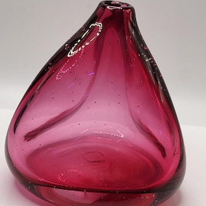 May include: A teardrop-shaped, ruby-coloured glass vase. The vase has a wide, open base that tapers to a narrow opening at the top. Small air bubbles are visible throughout the glass. The vase is a vibrant shade of red.