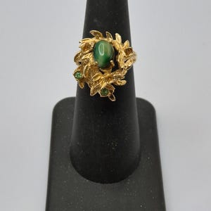 May include: A gold-toned ring featuring a central oval green gemstone, surrounded by a floral design with smaller green accent stones. The ring is displayed on a black ring holder against a white background.
