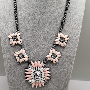 May include: A statement necklace with a dark gunmetal chain and a floral pendant. The pendant has pink and white petals with clear crystal accents. The necklace is displayed on a grey mannequin.
