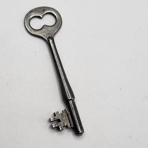 May include: A vintage metal key with a figure-eight shaped handle. The key has a long, slender stem and a detailed bit. The key is silver-toned and appears to be made of steel.