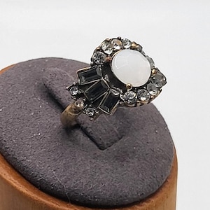 Könnte beinhalten: Ein Ring im Vintage-Stil mit einem zentralen, runden, weißen Edelstein, umgeben von klaren und schwarzen Akzentsteinen. Der Ring hat ein goldfarbenes Band und wird auf einem grauen, filzbeschichteten Ständer präsentiert.
