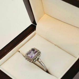May include: A silver ring featuring a rectangular, light purple amethyst gemstone set in a textured, rope-style metal border. The ring is displayed inside a dark brown wooden jewelry box with a cream-colored interior lining and a hinged lid.