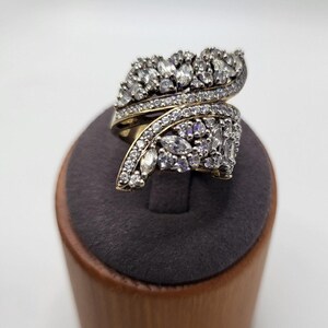 May include: An ornate, gold-toned ring with a swirling design. The ring is encrusted with numerous clear and marquise-cut gemstones. The ring is displayed on a wooden stand with a dark gray top, against a white background.