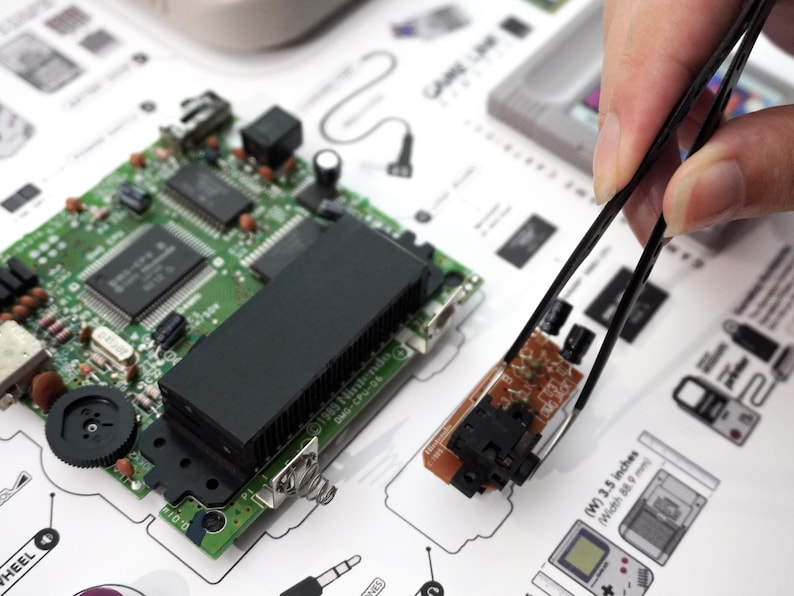 Close-up of tweezers arranging Game Boy circuit boards and internal components on a custom teardown art layout – arte tecnológico, zerlegte Konsole, retro console wandkunst.