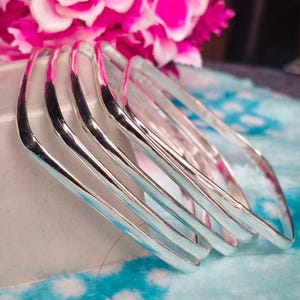 May include: A stack of five silver-colored, square-shaped bangles. The bangles are stacked on a white container, with a backdrop of pink flowers and a blue and white patterned surface.
