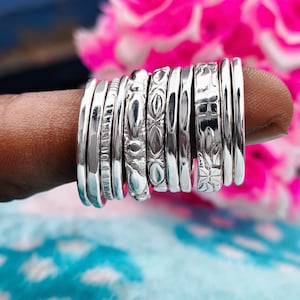 Op de afbeelding: Een verzameling zilveren ringen, elk met een uniek ontwerp, gedragen op een vinger. De ringen variëren in breedte en hebben patronen zoals kralenranden, bloemmotieven en gladde banden. De achtergrond bevat roze bloemen en een blauw-wit patroon.