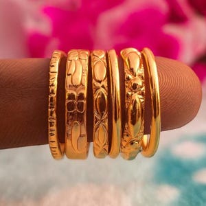 Pode incluir: Seis anéis dourados com designs variados, incluindo motivos florais e geométricos. Os anéis são exibidos em um dedo. O fundo está desfocado, com tons rosa e azul. Os designs dos anéis incluem faixas texturizadas e motivos florais.