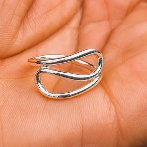 Könnte beinhalten: Ein silberfarbener Ring mit einem Doppelschlaufendesign. Der Ring besteht aus einem glänzenden, reflektierenden Metall und hat eine glatte Oberfläche. Der Ring wird auf einem hellorangen Hintergrund gezeigt.