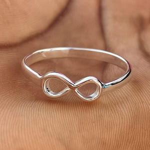 Op de afbeelding: Een zilverkleurige ring met een oneindigheidssymbool. De ring heeft een gladde, gepolijste afwerking en een eenvoudige band. Het oneindigheidssymbool staat centraal. De ring wordt getoond tegen een warme, neutrale achtergrond.