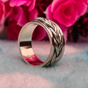 Peut inclure: Une bague argentée avec un motif tressé sur la bande extérieure. La bague est mise en valeur sur un fond de fleurs roses et une surface bleu clair. L'intérieur de la bague est argenté poli.