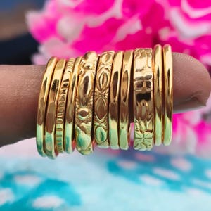 May include: A selection of gold-coloured rings, each featuring a distinct design, such as etched patterns and plain bands. The rings are stacked together, highlighting their diverse textures and styles. The rings are set against a blurred backdrop of pink and blue.