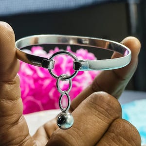 May include: A silver bangle bracelet with a circular charm and a dangling sphere. The bracelet is held up against a blurred background of pink flowers. The bracelet is made of polished metal.