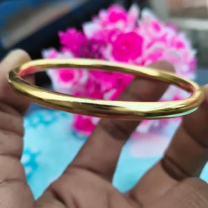 Puede incluir: Una pulsera de brazalete de color dorado. La banda lisa y redondeada refleja la luz, creando una apariencia brillante y brillante. La pulsera se sostiene en una mano, con un fondo borroso de flores rosas y blancas.