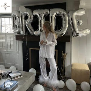 May include: A white satin pajama set with feather trim. Silver foil balloons spelling "BRIDE" are suspended above a fireplace. White balloons are scattered around the room, creating a celebratory atmosphere. The pajama set is a classic design.