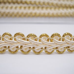 Puede incluir: Borde blanco y dorado con un diseño tejido decorativo. El borde tiene un patrón elevado y en bucle con acentos dorados.