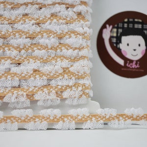 Op de afbeelding: Witte en gouden gerimpelde trim met een delicaat kantpatroon. De trim wordt getoond in een rol en een close-up van het detail.