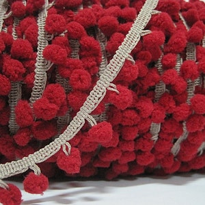 May include: Red and beige pom-pom trim with a woven texture. The trim is made of soft, fluffy pom-poms and a woven ribbon.