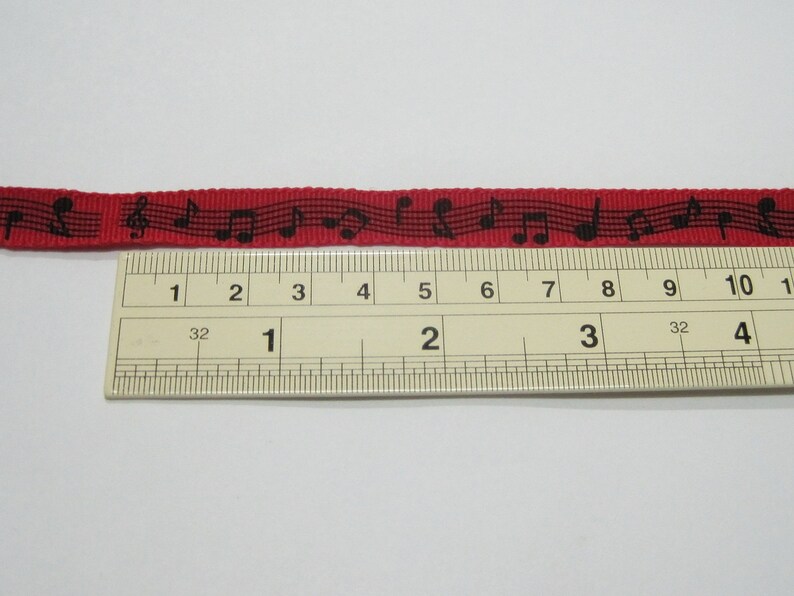 May include: Red ribbon with black musical notes printed on it. The ribbon is measured against a ruler, showing it is approximately 4 inches long.