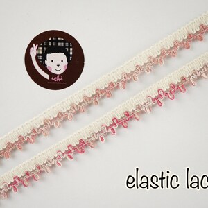 Puede incluir: Borde de encaje elástico blanco con flecos rosa y blanco. El encaje está doblado por la mitad y los flecos cuelgan. El texto "elastic lace" está escrito debajo del encaje.