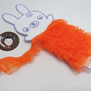 May include: Orange ruffled trim with a sheer, slightly sparkly texture. The trim is folded and displayed on a white background.