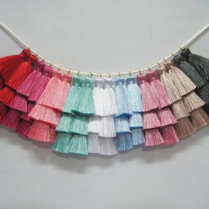 Puede incluir: Una guirnalda de 12 borlas multicolores, cada borla está hecha de tres capas de flecos. Las borlas están dispuestas en un patrón de arcoíris, comenzando con rojo y terminando con gris. Las borlas están enhebradas en un cordón blanco.