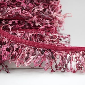 May include: Close-up of a burgundy trim with a deep pink and white fringe. The trim is made of a soft, textured fabric.