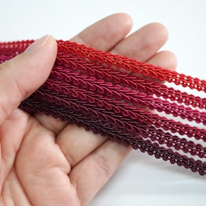 May include: Close-up of several strands of red and burgundy decorative trim. The trim has a scalloped edge and is held in a hand. The colors range from a bright red to a deep burgundy.