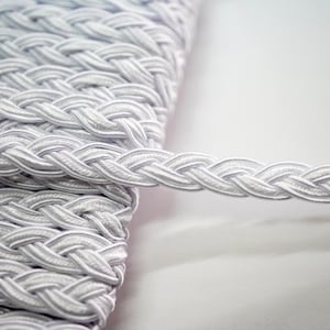May include: White braided trim with a woven texture. The trim is coiled and a single strand is extended.