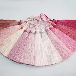Könnte beinhalten: Ein Set aus zehn rosa und weißen Seidenquasten, die in Fächerform angeordnet sind. Die Quasten sind in verschiedenen Rosatönen gehalten, von einem tiefen Rot bis zu einem zarten Blush.