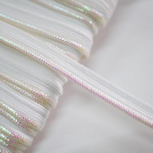 May include: Close-up of a spool of white elastic ribbon with iridescent, shimmering accents. The ribbon is tightly wound, with a single strand extending outwards. The material appears to be a soft, flexible fabric, suitable for various crafting projects.