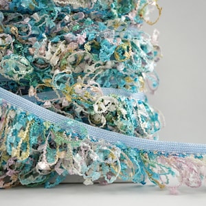 May include: A close-up of a spool of light blue and turquoise fringe trim. The fringe is made of a variety of colors, including pink, white, and gold.