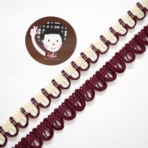 May include: Two decorative ribbons with a scalloped edge. One ribbon is cream and dark brown, the other is burgundy. A circular logo with a cartoon character and the word "ichi" is in the upper left corner.