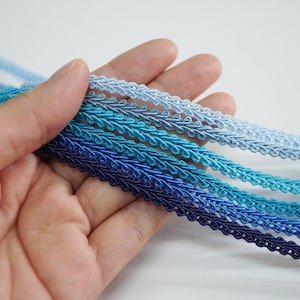 May include: A hand holds several strands of blue decorative trim. The trim is a woven design with a scalloped edge. The colors range from light to dark blue, creating a gradient effect. The trim is likely used for sewing or crafting.