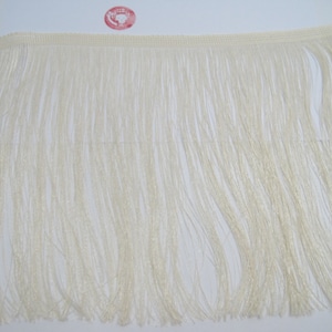 Puede incluir: Primer plano de un ribete de flecos blanco. El fleco está hecho de hilos finos y sedosos que miden aproximadamente 2,5 cm de largo.