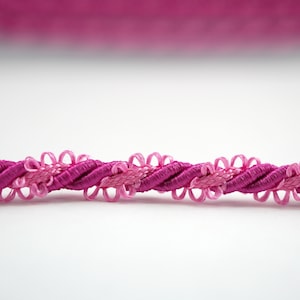May include: A close-up of pink twisted cord trim with a decorative flower design. The cord is made of a soft, silky material and has a subtle sheen.