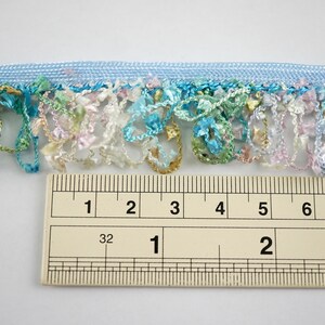 May include: A close-up of a light blue trim with a fringe of colorful, multi-colored threads. The trim is measured with a ruler, showing it is 2 inches wide.