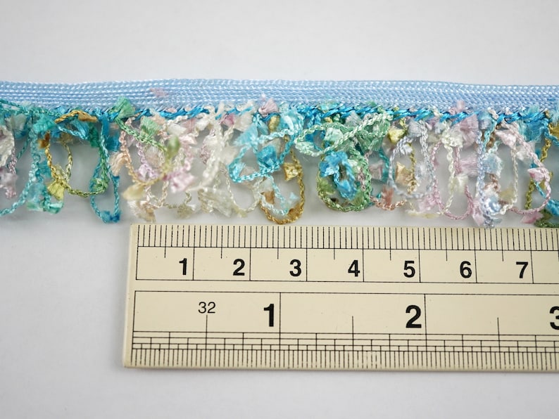 May include: A close-up of a light blue trim with a fringe of colorful, multi-colored threads. The trim is measured with a ruler, showing it is 2 inches wide.