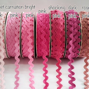 May include: A collection of pink ric rac ribbon spools in various shades, from light pink to dark rose. The spools are labeled with color names like "sweet pink" and "shocking pink". The ribbon has a wavy, decorative edge.