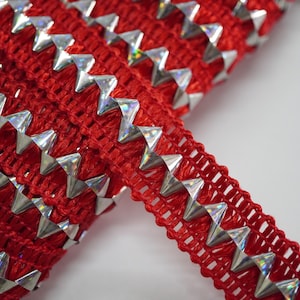 May include: Close-up of red decorative trim with silver, triangular studs. The trim has a woven texture and the studs are arranged in a zig-zag pattern. The studs have a reflective, iridescent finish, adding a touch of sparkle.