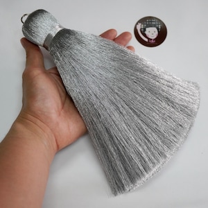 Puede incluir: Una borla grande de color gris plateado con un lazo en la parte superior. La borla se sostiene en una mano, mostrando su textura suave y fluida. La borla probablemente se utiliza con fines decorativos.